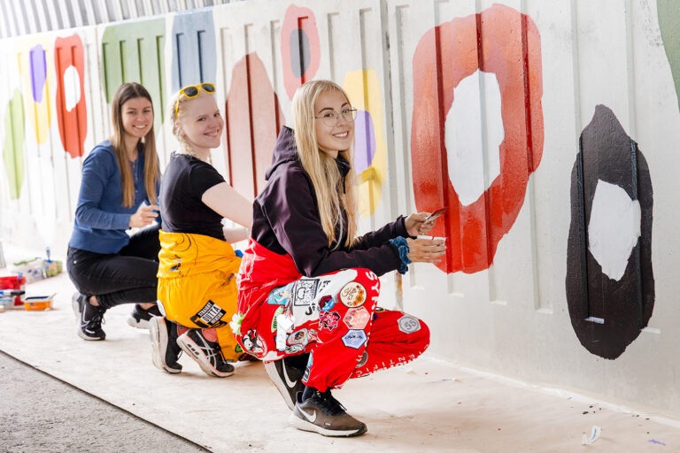 Kolme iloista, haalaripukuista opiskelijaa maalaa alikulkutunnelin seinään värikkäitä kuvioita.
