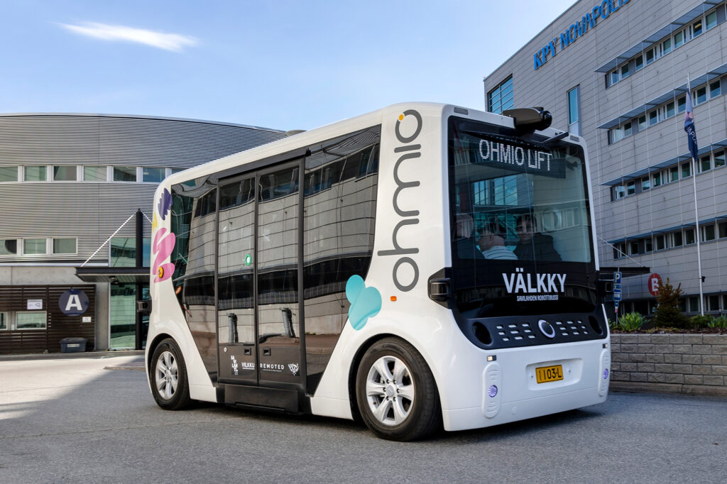 Pieni, valkoinen robottibussi, jonka kyljessä on erivärisiä muotoja, on pysäköity parkkipaikalle.