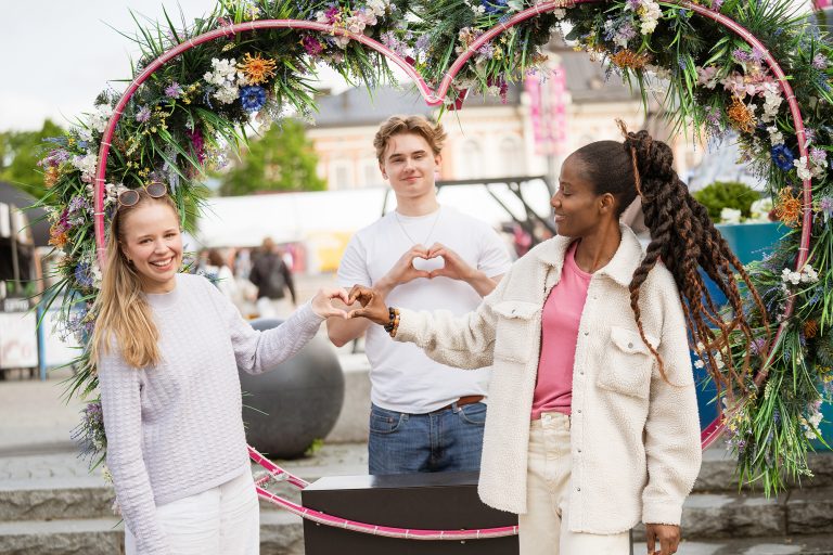 Kolme nuorta torilla sydämenmuotoisen kukkaköynnöksen edessä.