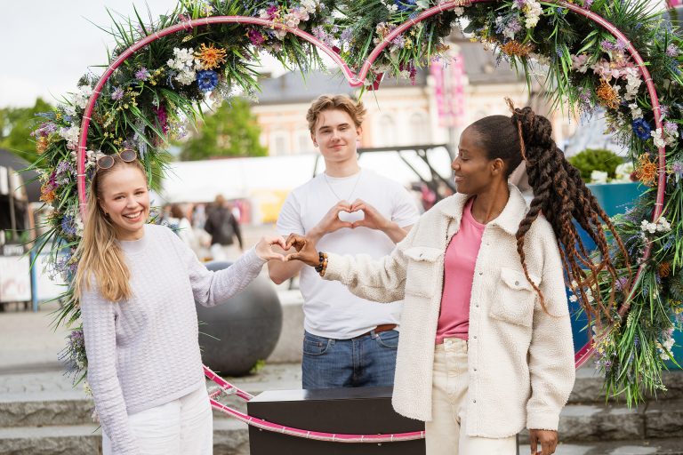Kolme nuorta hymyilee torilla sydämenmuotoisen kukkatelineen edessä.